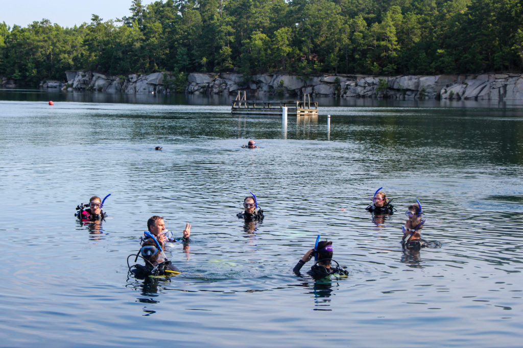 A group of divers are floating at the surface of the rock quarry diving park. A dive leader is reviewing the typical dive signs.