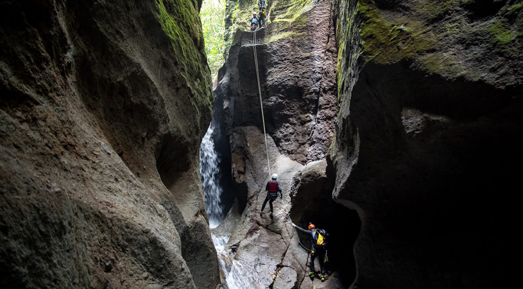 Two cliff faces are on either side, with a crevice down the center. Climbers are coming down ropes in the center with a waterfall behind them.