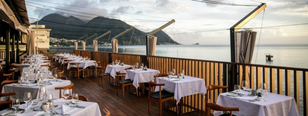 The dining room at Fort Young in Dominica, the tables are set up in groups of 4 and overlooks the ocean to the right. Mountains can be seen in the background. 