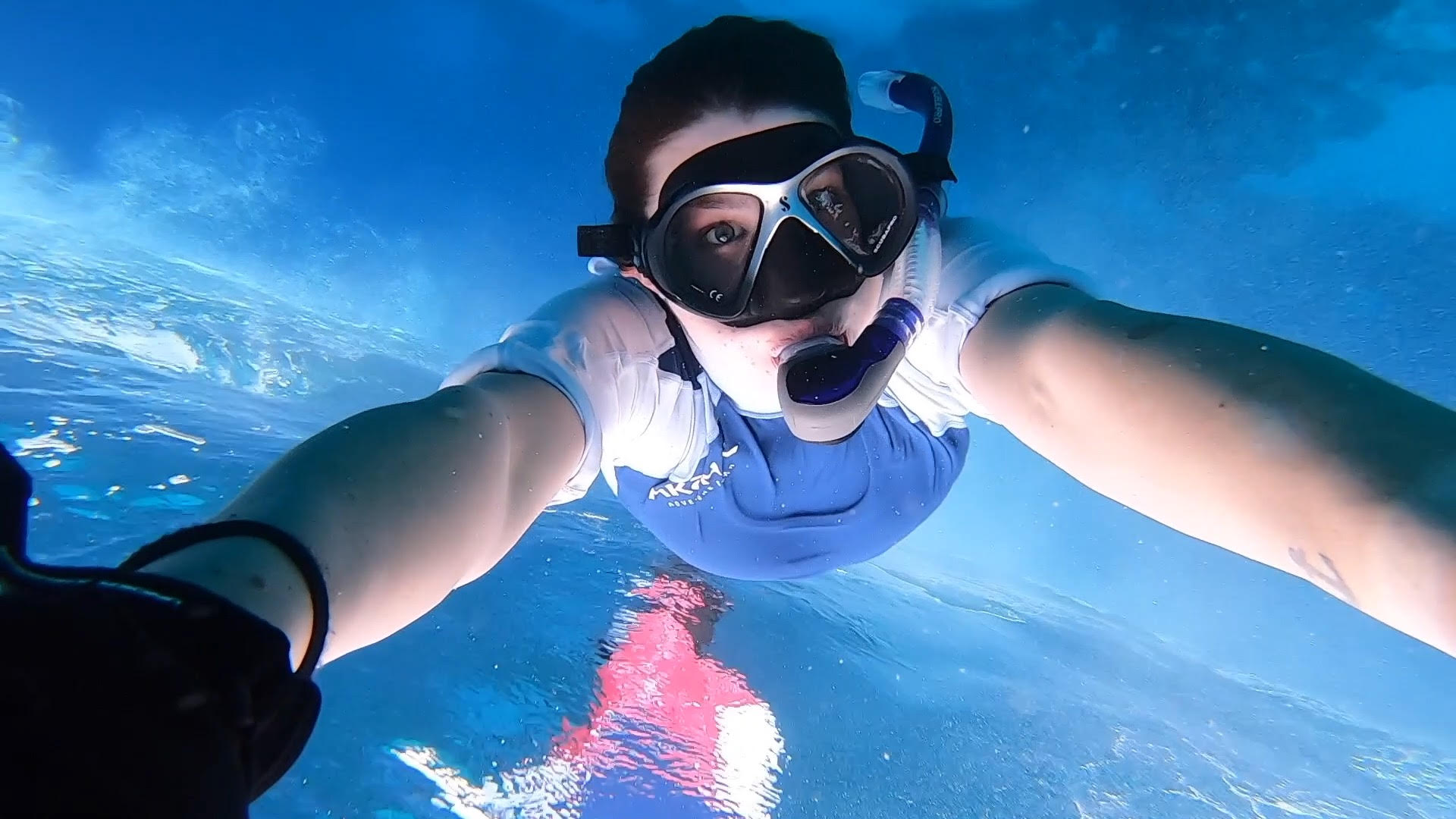 A young woman holds on to something off screen, she's spun underwater while wearing a mask and snorkel. 