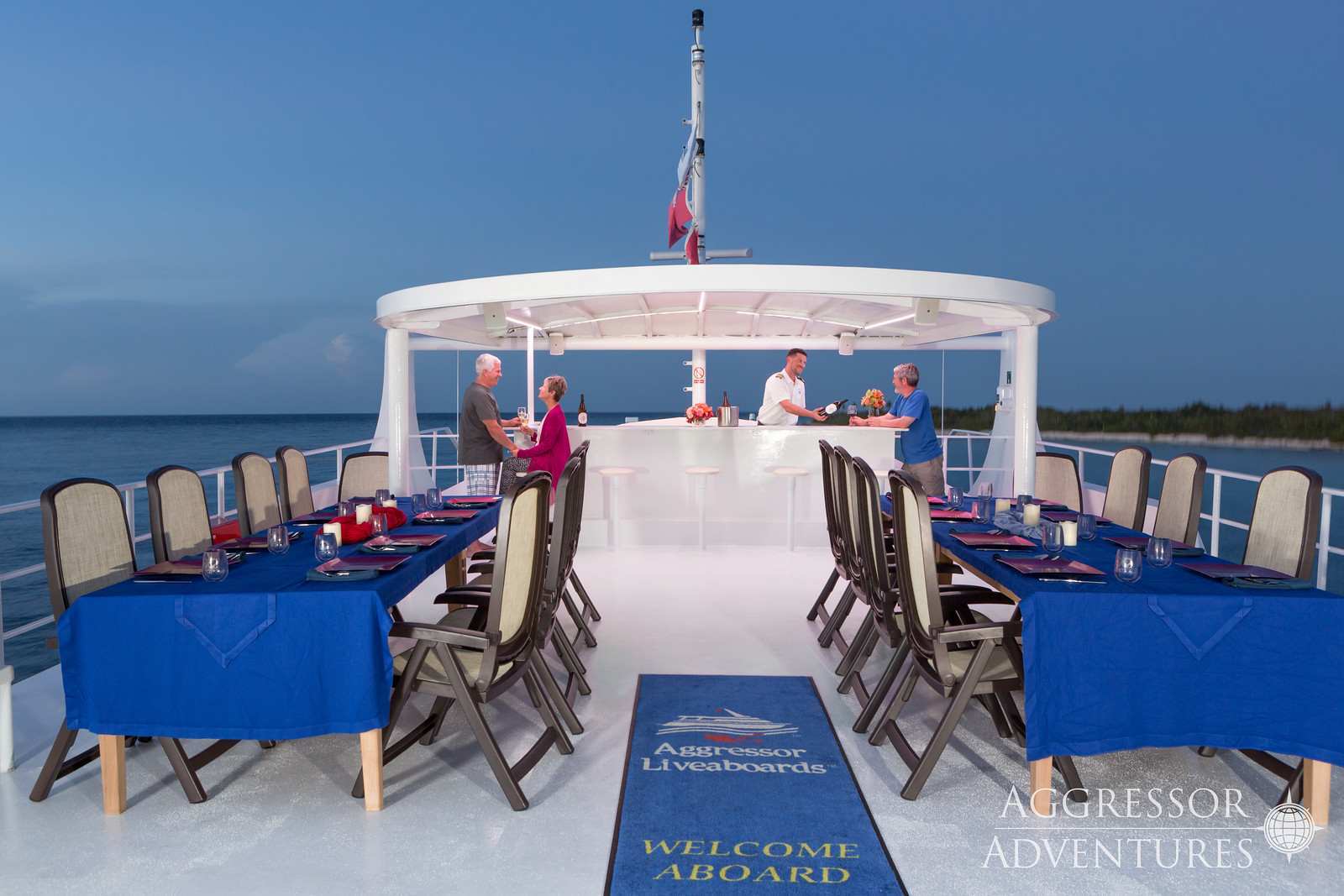 On top of the Cayman Aggressor V, there are several tables laid out with chair and table cloths. In the back, there is a bartender serving a couple at a bar.