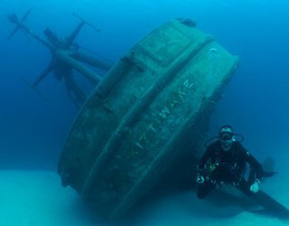 A diver floats at the bottom of the ocean underneath a piece of a wrecked ship. 