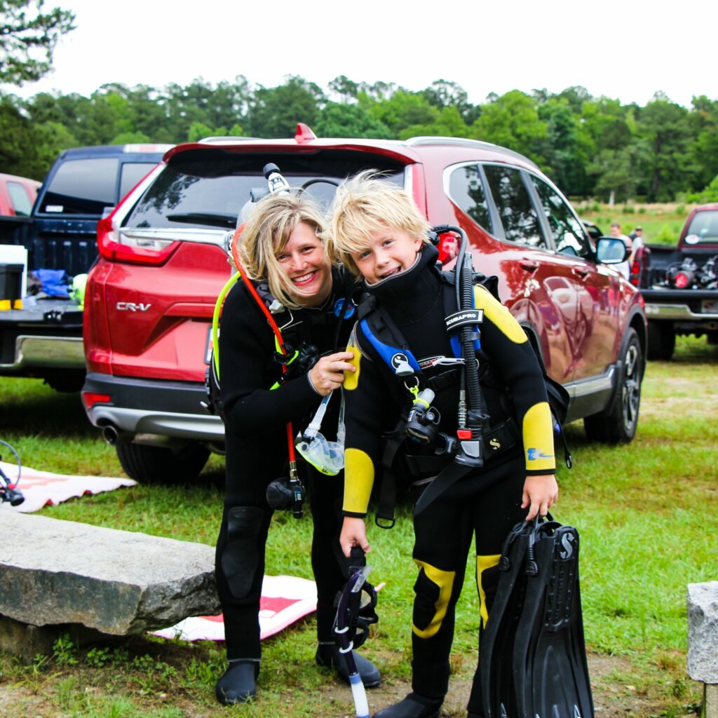 Two divers are standing beside each other smiling at the camera. The mother is on the left, bending over to stand head to head with her son, who is on the right.