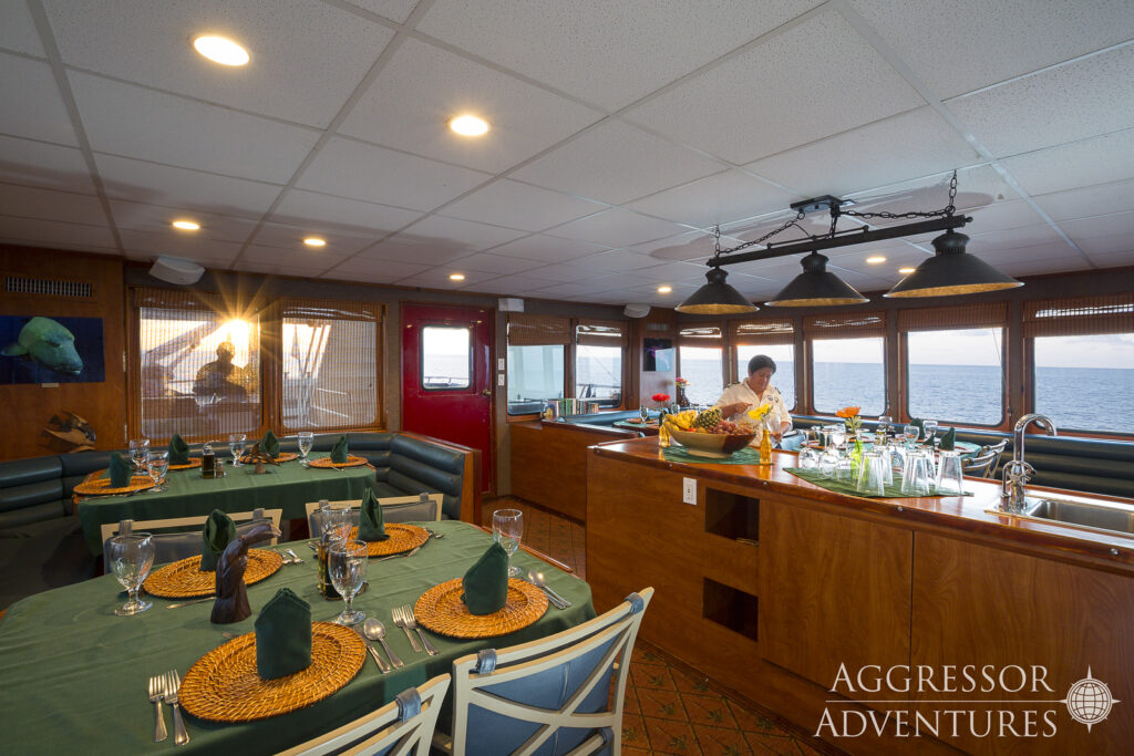 The dining room, or restaurant, aboard the Belize Aggressor IV has green tables adorned with decorations and place settings already in place. In the middle of the room, there's a buffet table for food service. 